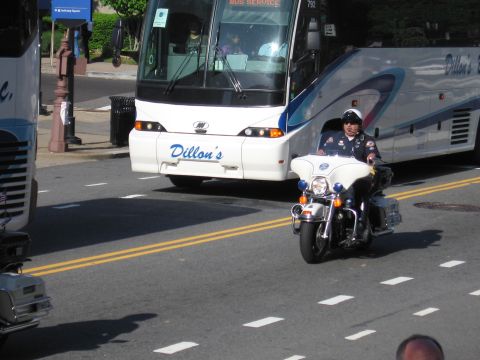 1991 Ford LTD Crown Victoria - lone helmeted motorcycle officer rides past the front of a white Dillon's Bus Service motorcoach on a sunny street.