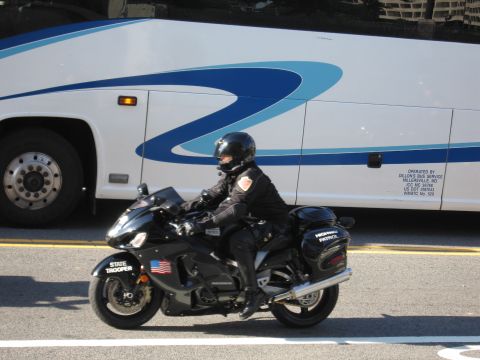 1991 Ford LTD Crown Victoria - state trooper in black gear rides a black sport motorcycle marked STATE TROOPER past a white-and-blue motorcoach.