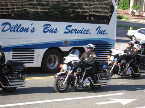1991 Ford LTD Crown Victoria - Police motorcycle officers ride past a white-and-blue Dillon's Bus Service motorcoach on a sunlit city street.