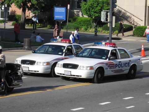 1991 Ford LTD Crown Victoria - Two white Ford Crown Victoria cruisers marked POLICE sit angled on a city street with pedestrians and a building behind.