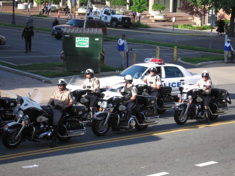 1991 Ford LTD Crown Victoria - row of sheriff's deputies on motorcycles lines up in the street beside a white police car near a dumpster.