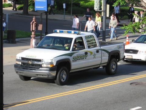 1991 Ford LTD Crown Victoria - white-and-green Chevrolet Silverado crew-cab pickup marked SHERIFF drives a city street as pedestrians cross behind it.