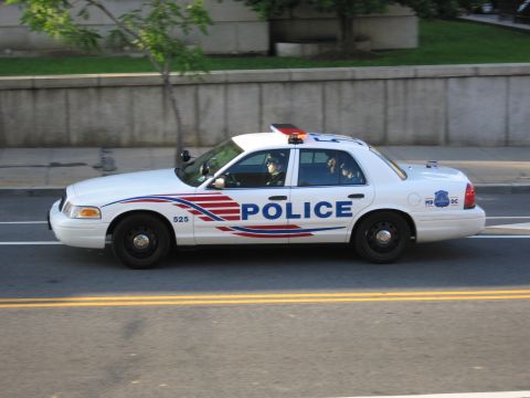 1991 Ford LTD Crown Victoria - white Ford Crown Victoria marked POLICE drives along an urban roadway past a concrete retaining wall.
