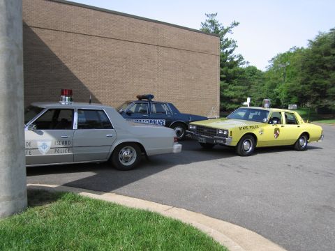 1991 Ford LTD Crown Victoria - silver Rhode Island State Police sedan, a yellow State Police sedan, and a dark cruiser are parked beside a brick building.