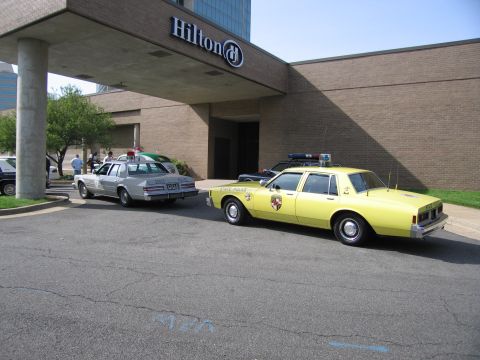 1991 Ford LTD Crown Victoria - yellow vintage State Police sedan and a silver sedan are parked under the entrance canopy of a Hilton hotel.