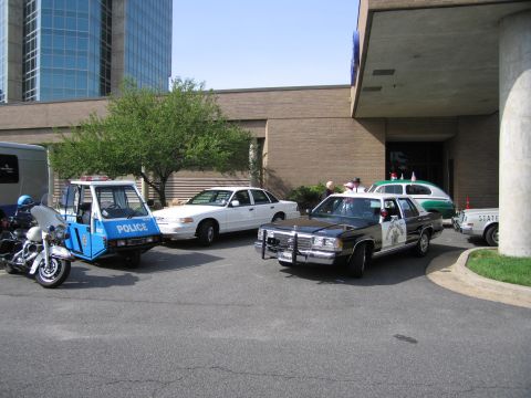 1991 Ford LTD Crown Victoria - Assorted police vehicles, including a three-wheeled POLICE motorcycle and vintage sedans, are gathered beside a brick building.