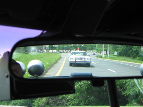 1991 Ford LTD Crown Victoria - Seen through a vehicle's interior rear-view mirror, a white police car follows on a tree-lined suburban road.