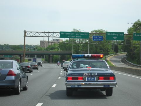 1991 Ford LTD Crown Victoria - rear of a dark blue Chevrolet Caprice with a lightbar and EMER-911 lettering is seen on a highway beneath green overhead signs.