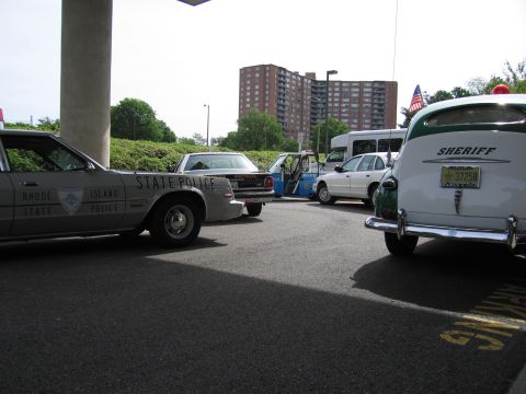 1991 Ford LTD Crown Victoria - black-and-white Rhode Island State Police sedan and a green-and-white Sheriff car are parked outdoors with a tall apartment behind.