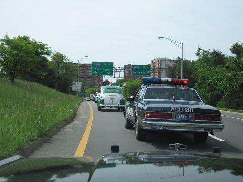 1991 Ford LTD Crown Victoria - dark Chevrolet Caprice police car follows a white antique Sheriff car along a highway shoulder beneath green overhead signs.