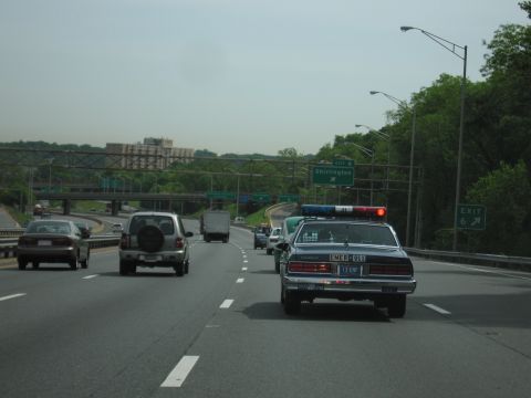 1991 Ford LTD Crown Victoria - dark blue Chevrolet Caprice fitted with a police-style lightbar travels a multilane highway past overhead green exit signs.