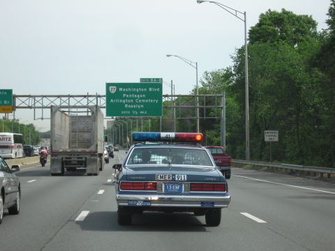 1991 Ford LTD Crown Victoria - dark blue Chevrolet Caprice police car drives a highway next to a box truck, below a green sign for Washington Blvd and the Pentagon.
