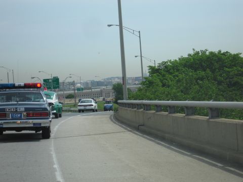1991 Ford LTD Crown Victoria - black-and-white police car with light bar follows cars down an elevated highway curve, with a hazy city skyline and guardrail beyond.