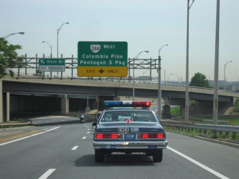1991 Ford LTD Crown Victoria - Chevrolet Caprice police cruiser drives a multi-lane highway beneath a green overhead sign reading Route 244 West, Columbia Pike, Pentagon…