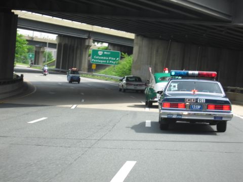 1991 Ford LTD Crown Victoria - dark police car with red and blue light bar rounds a curving roadway under a concrete overpass, trailing other vehicles in the distance.