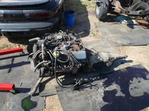 1975 Ford Ranchero - A removed engine and transmission assembly sits on a black mat on the ground, with a black Pontiac Firebird in the background. A blue bucke…