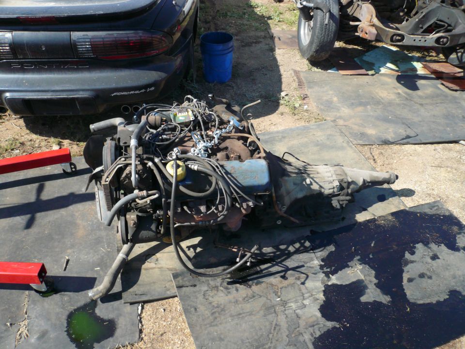 Engine and transmission assembly on a mat with a black Pontiac Firebird in the background.