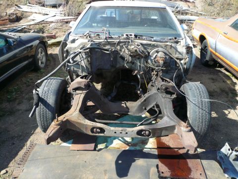 1975 Ford Ranchero - The front end of a disassembled vehicle is shown, with the engine bay and chassis exposed. A black car and an orange car are partially visi…