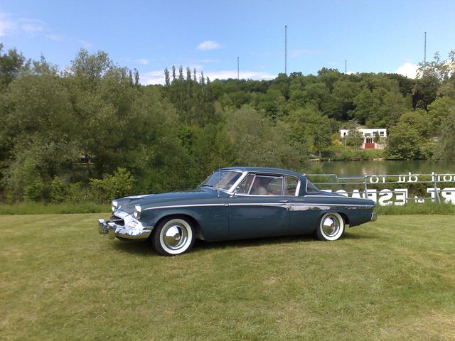 Blue 1955 Studebaker coupe with white-wall tires parked on grass near a lake