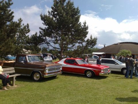1991 Ford Crown Victoria - A brown pickup truck and a red and white coupe are parked on a grassy field with people in the background.
