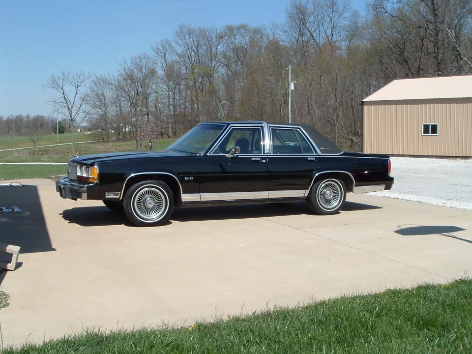 Black Ford LTD Crown Victoria sedan side view on driveway near a tan pole barn