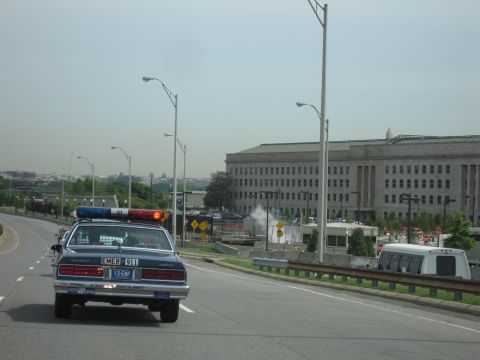 1991 Ford LTD Crown Victoria - Chevrolet police cruiser travels a divided roadway with the large Pentagon building and a parked RV visible to the right.