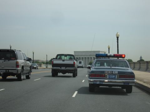 1991 Ford LTD Crown Victoria - Chevrolet Caprice police cruiser and a white pickup cross a bridge with the columned Lincoln Memorial visible ahead through the haze.