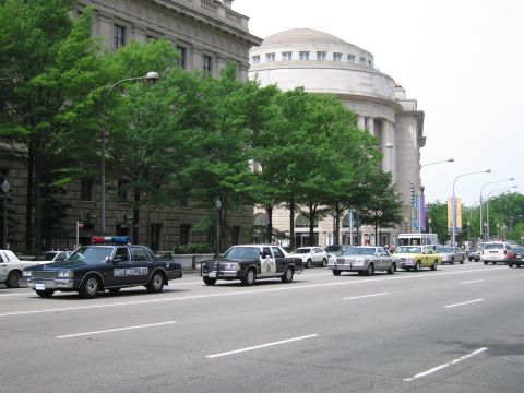 1991 Ford LTD Crown Victoria - Black-and-tan and other vintage police cars sit on a city street beside a large domed classical stone building and green trees.
