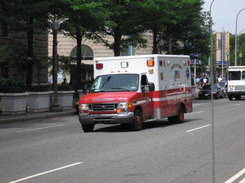 1991 Ford LTD Crown Victoria - red-and-white Ford box-body ambulance marked District of Columbia drives a tree-lined city street with a truck and parked car nearby.