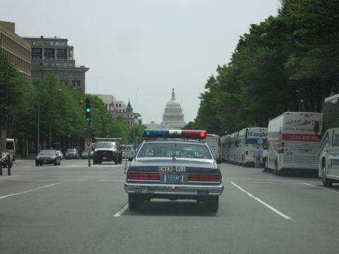 1991 Ford LTD Crown Victoria - Chevrolet police cruiser drives a wide city avenue lined with buses and trucks, the U.S. Capitol dome framed in the distance.