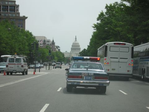 1991 Ford LTD Crown Victoria - Rear view of a Chevrolet police cruiser on a city street flanked by a tour bus and a van, the U.S. Capitol dome rising at the end of the av…