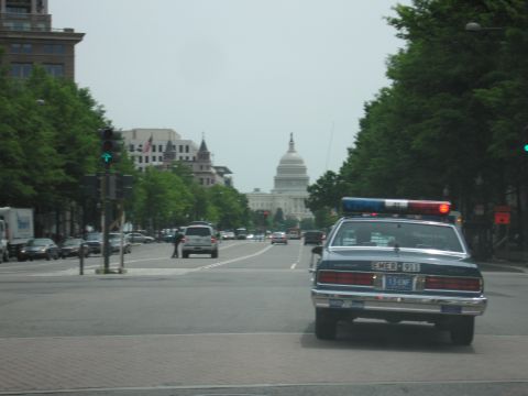 1991 Ford LTD Crown Victoria - Chevrolet police cruiser sits at a brick-paved intersection on a tree-lined avenue with the U.S. Capitol dome visible far ahead.