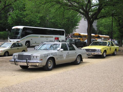1991 Ford LTD Crown Victoria - silver State Police sedan with push bar parks beside a yellow taxi on a gravel lot, with tour buses and yellow school buses in the backgrou…