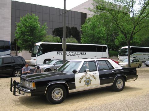 1991 Ford LTD Crown Victoria - black-and-white Highway Patrol Ford sedan with star emblem is parked on gravel in front of a Wise Coaches tour bus and a dark building.