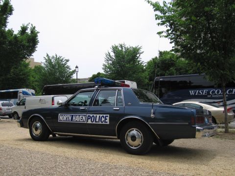 1991 Ford LTD Crown Victoria - Side profile of a dark blue North Haven Police Chevrolet Caprice cruiser parked on a gravel lot among vans and tour buses.