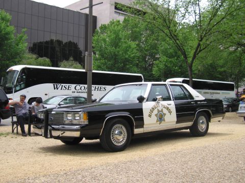 1991 Ford LTD Crown Victoria - black-and-white Highway Patrol Ford LTD Crown Victoria with push bar parks on gravel before Wise Coaches tour buses as two people sit nearb…