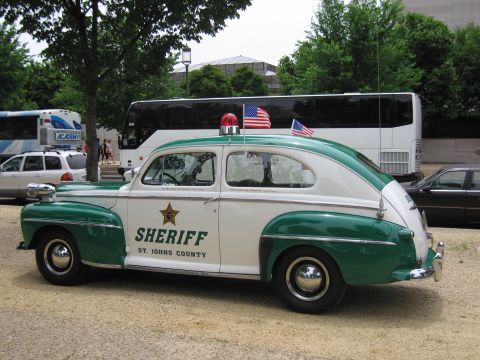 1991 Ford LTD Crown Victoria - restored green-and-white 1940s Ford sedan marked SHERIFF St. Johns County, with red beacon and two American flags, parked beside tour buses.