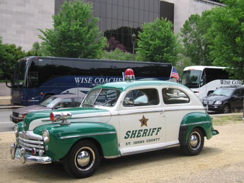 1991 Ford LTD Crown Victoria - Front quarter view of a restored green-and-white 1940s Ford St. Johns County Sheriff car with chrome grille and red beacon, buses behind.
