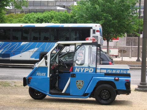 1991 Ford LTD Crown Victoria - blue-and-white three-wheeled NYPD police scooter with open cab and light bar is parked on gravel beside a transit bus and trees.