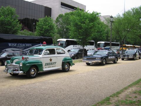 1991 Ford LTD Crown Victoria - restored vintage green-and-white sheriff's sedan leads a row of classic police cruisers parked along a gravel path near tour buses.