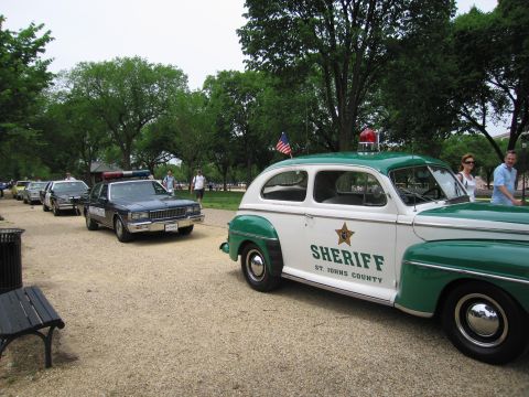1991 Ford LTD Crown Victoria - vintage green-and-white SHERIFF St Johns County sedan with light bar parked beside a dark sedan cruiser along a tree-lined gravel path.