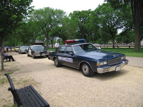 1991 Ford LTD Crown Victoria - dark blue State Police four-door sedan with roof light bar parked on a gravel path beside a wooden bench in a tree-lined park.