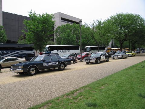1991 Ford LTD Crown Victoria - dark North Haven Police sedan with light bar leads a line of police cars and tour buses parked along a gravel path by a building.