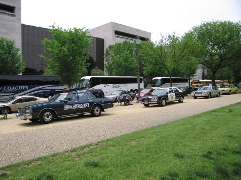 1991 Ford LTD Crown Victoria - North Haven Police sedan and other classic cruisers line a gravel path alongside Wise Coaches tour buses and a school bus.