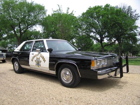 1991 Ford LTD Crown Victoria - black-and-white Highway Patrol four-door sedan with star emblem and push bar parked on a gravel path beneath shade trees.