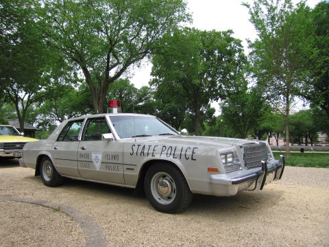 1991 Ford LTD Crown Victoria - silver Rhode Island State Police four-door sedan with red light bar and push bar parked on a gravel area among trees.