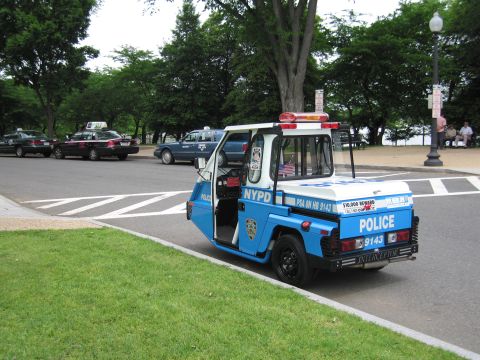 1991 Ford LTD Crown Victoria - small blue-and-white NYPD three-wheeled Interceptor police scooter with light bar parked on a roadway in a park setting.