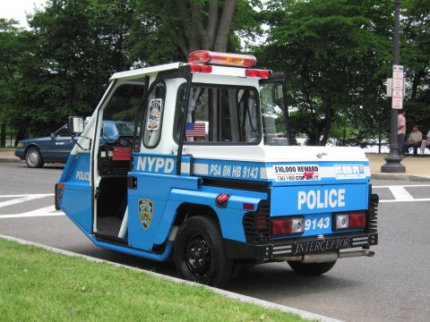 1991 Ford LTD Crown Victoria - blue-and-white NYPD Interceptor three-wheeled police scooter with open door and reward decals parked on a marked roadway.