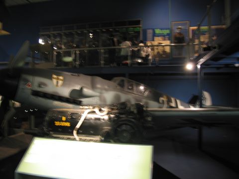 1991 Ford LTD Crown Victoria - gray German fighter aircraft with shield emblem displayed in a dim museum hall, with an exposed inline engine in the foreground (motion-blu…