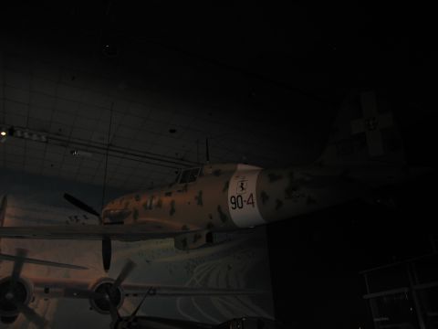1991 Ford LTD Crown Victoria - tan desert-camouflage fighter aircraft marked 90-4 with tail cross, dimly lit and suspended over a mural in a dark museum hall.